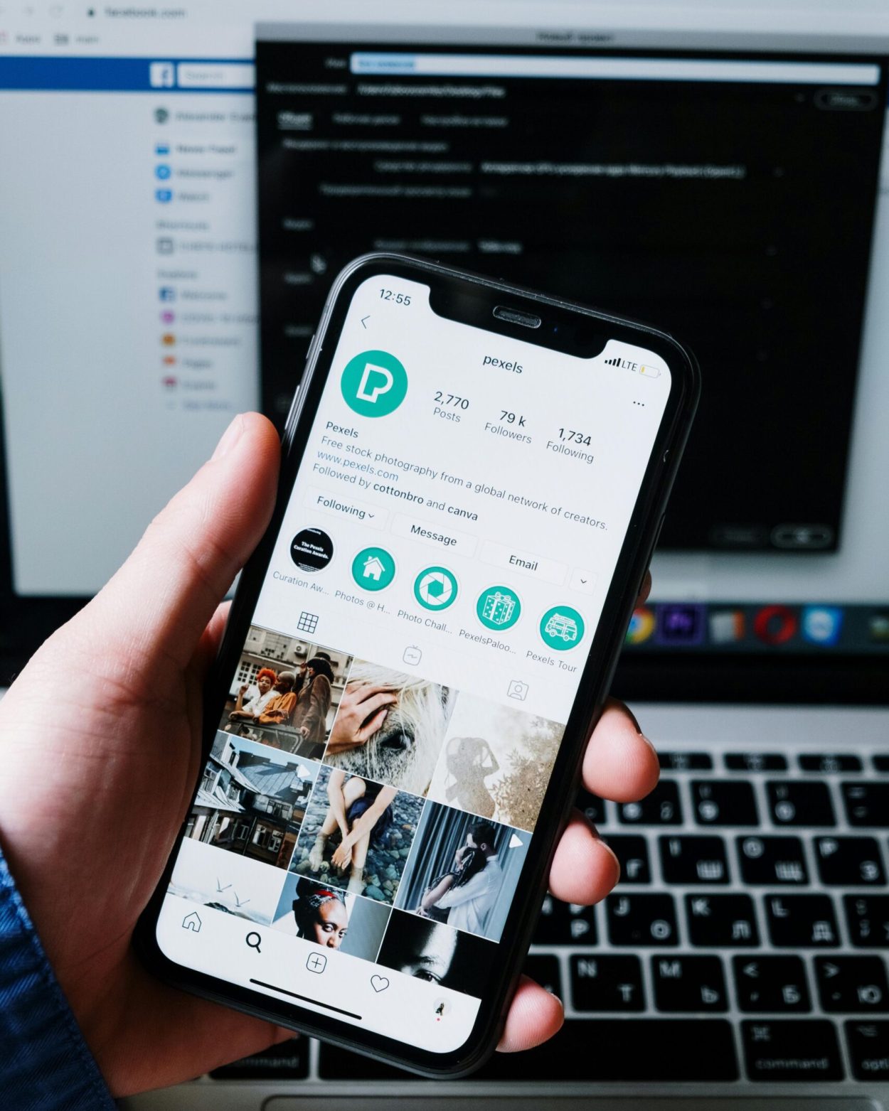 Hand holding smartphone displaying social media app with laptop in the background.